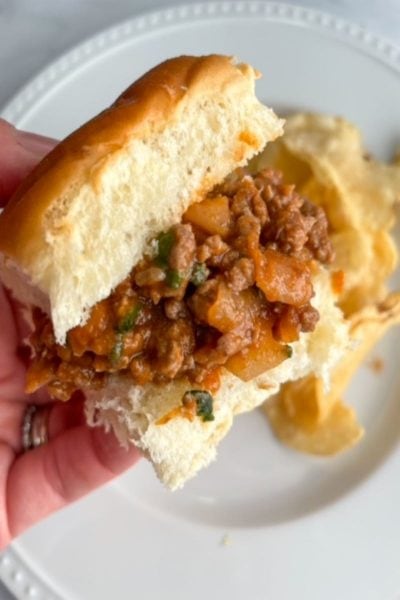 sloppy joe sandwich held above white plate with potato chips in background