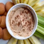 bowl of peanut butter dip sprinkled with cinnamon surrounded by cookies, celery and apple slices on white plate