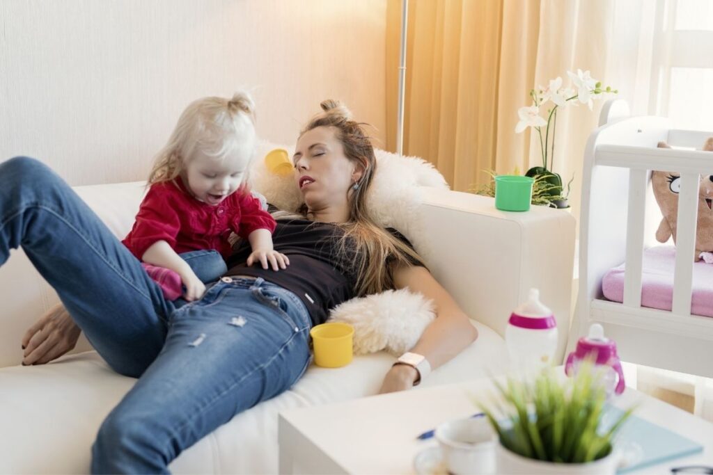Mom sprawled out exhausted on couch as toddler girl crawls across her